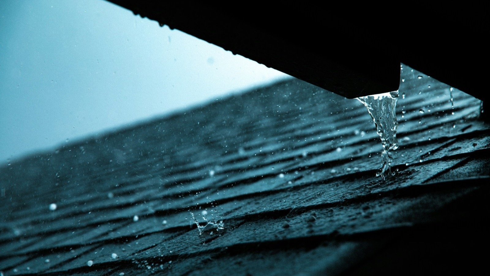 Water running off the edge of a dark shingle roof during heavy rain